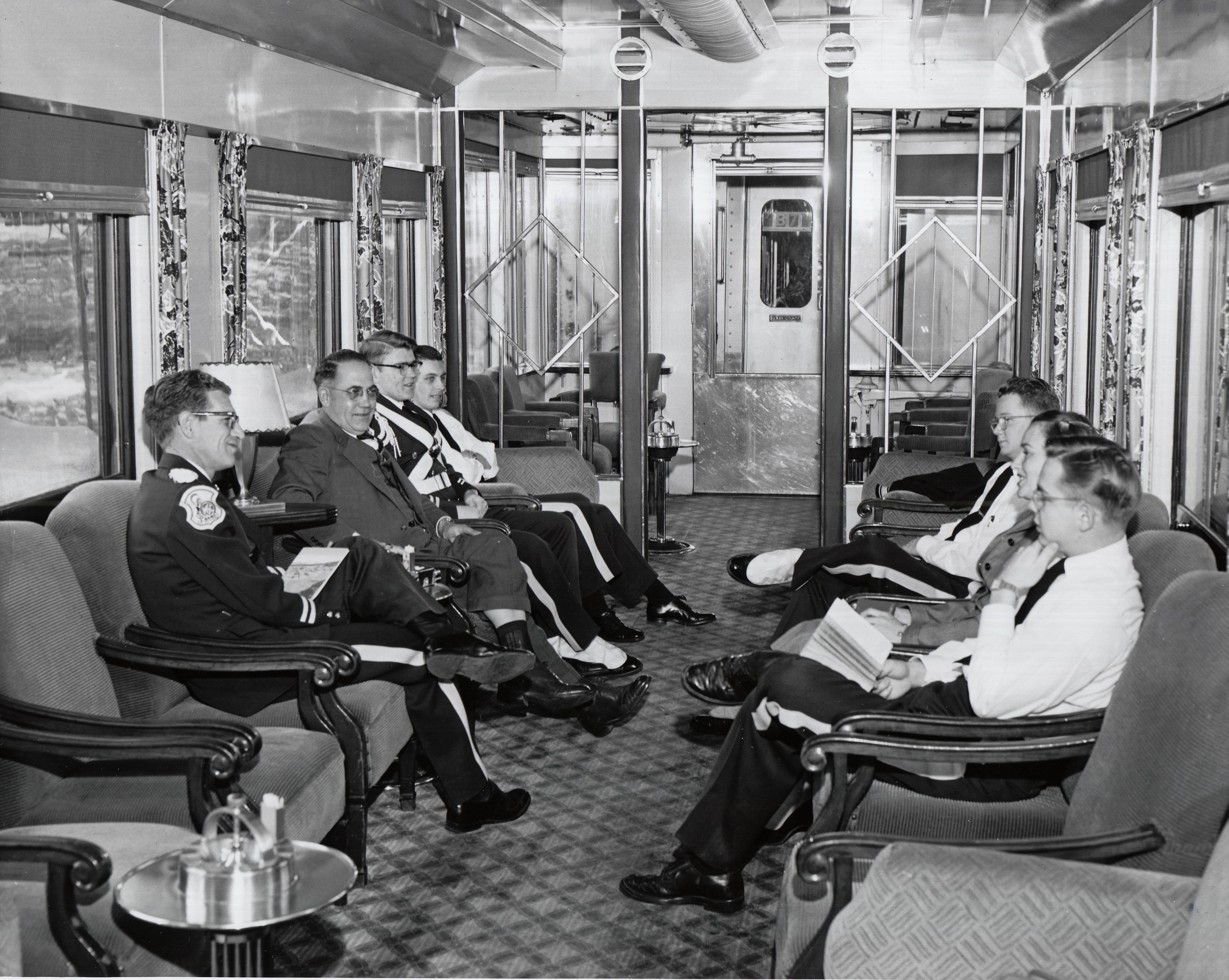 Seven people talk while seated in padded arm chairs in a railroad car