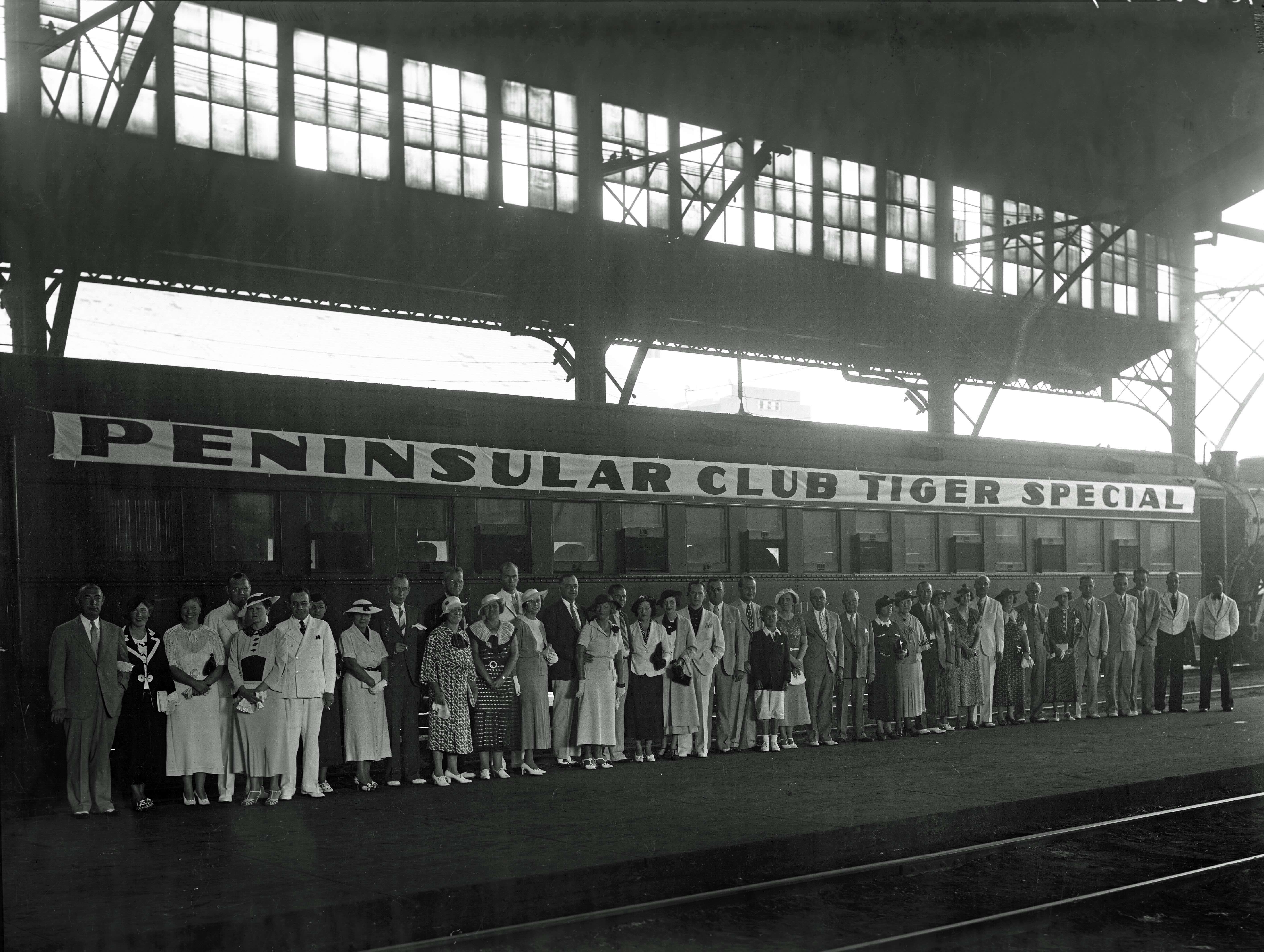 A line of people wait by a railroad track under a sign reading Peninsular Club Tiger Special