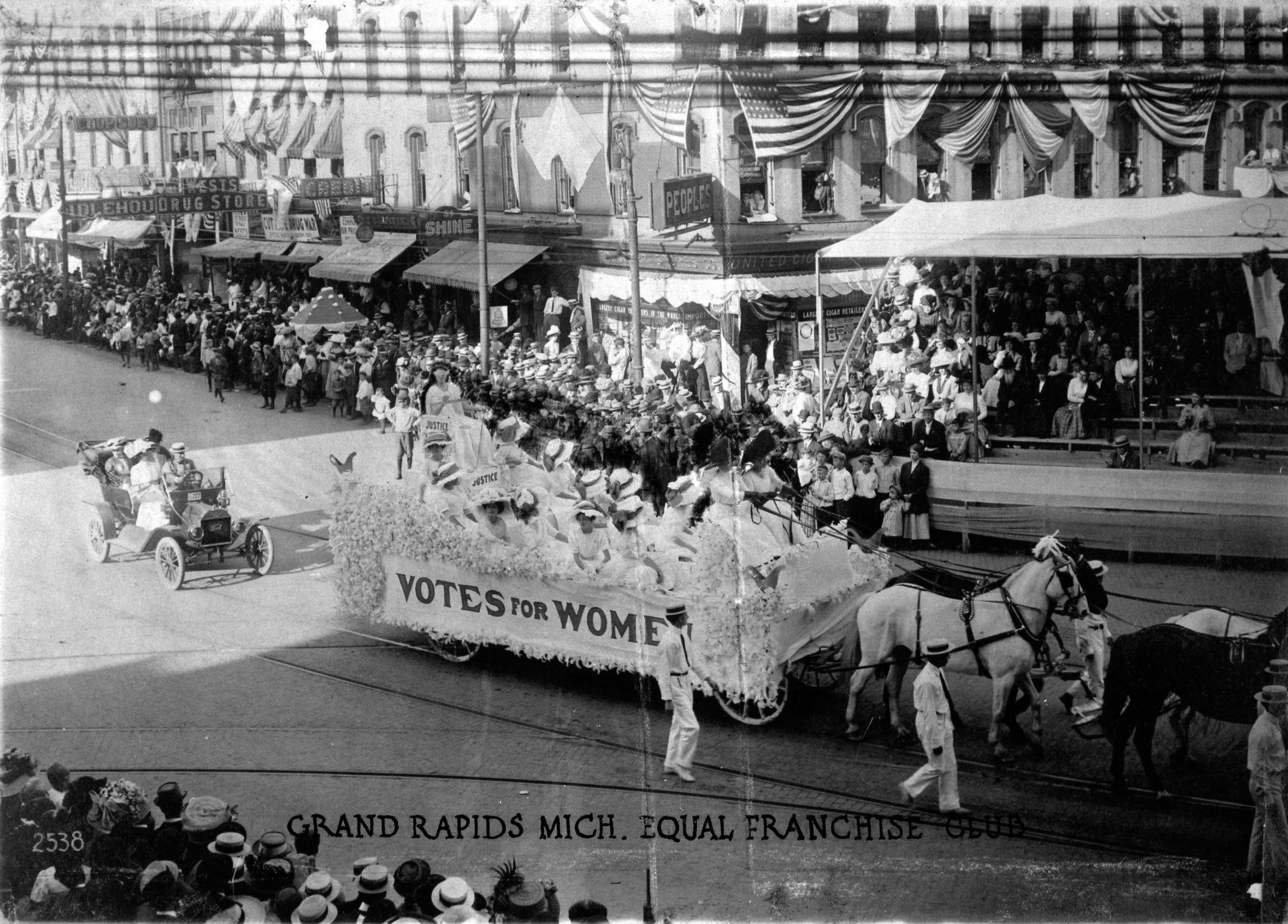 A wheeled float is pulled down a busy downtown street by a team of horses in 1910