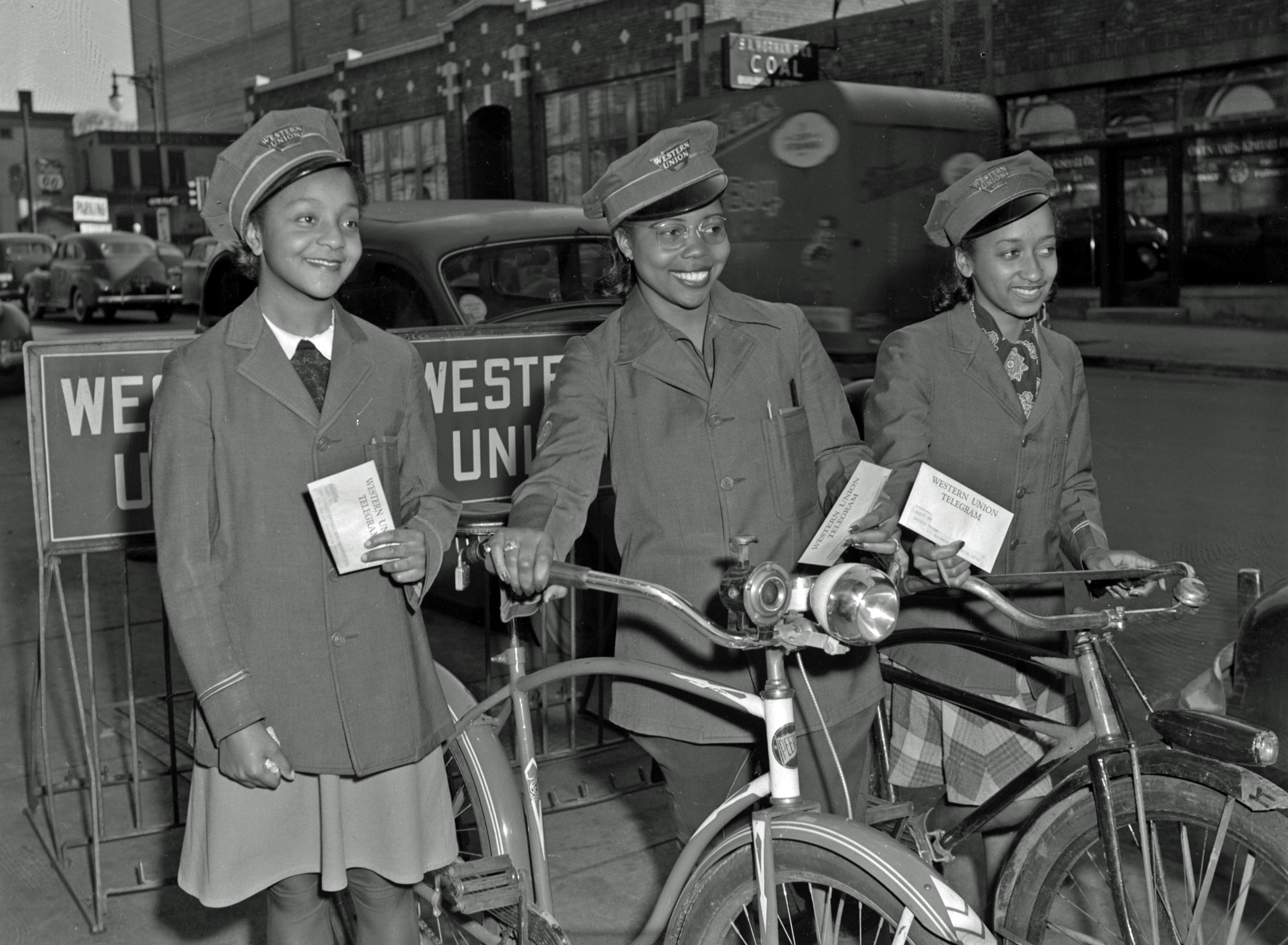 Three young African American women in Western Union uniforms smile and pose for a photo near bicycles outdoors while holding paper telegrams.