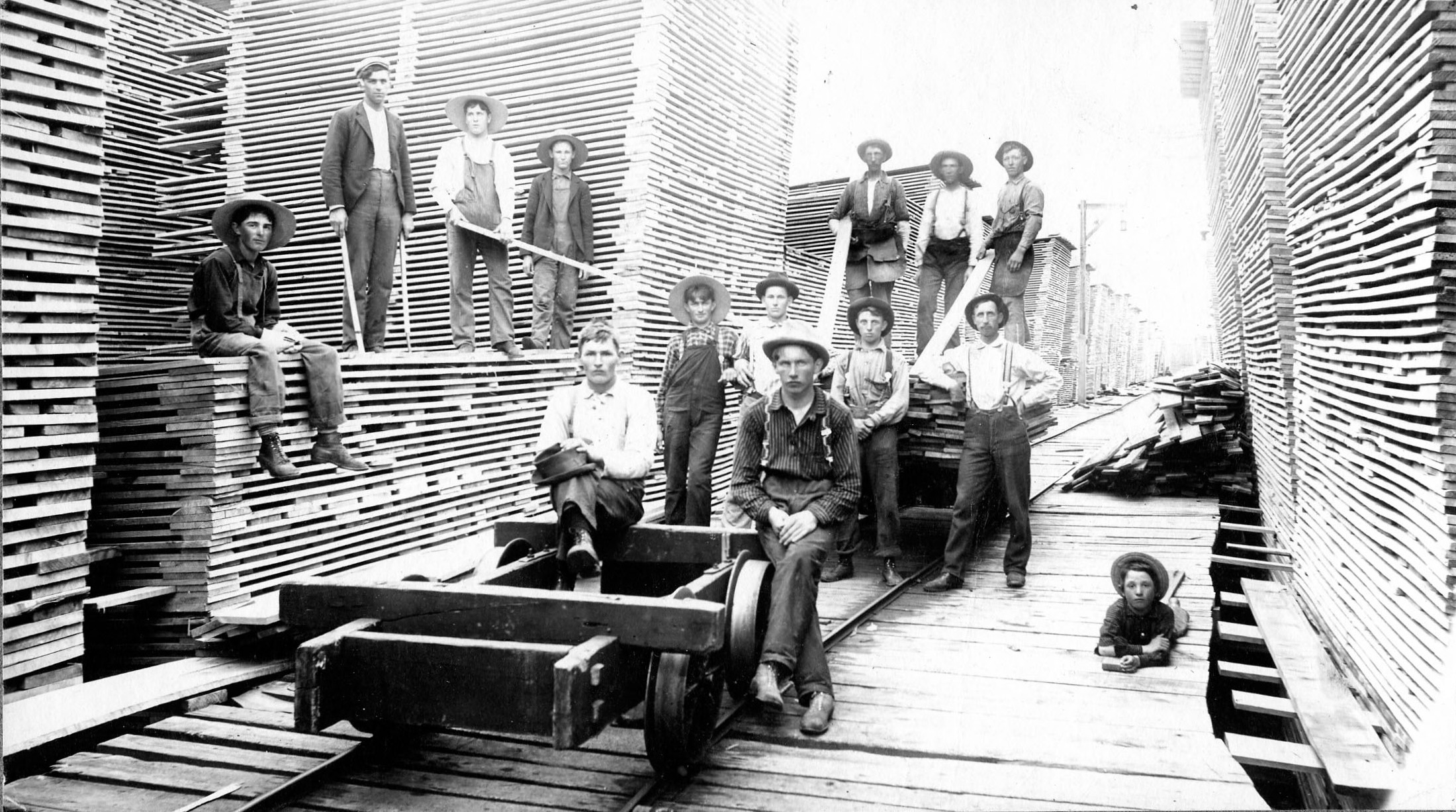 Several men sit or stand on a four wheel cart surrounded by tall piles of lumber