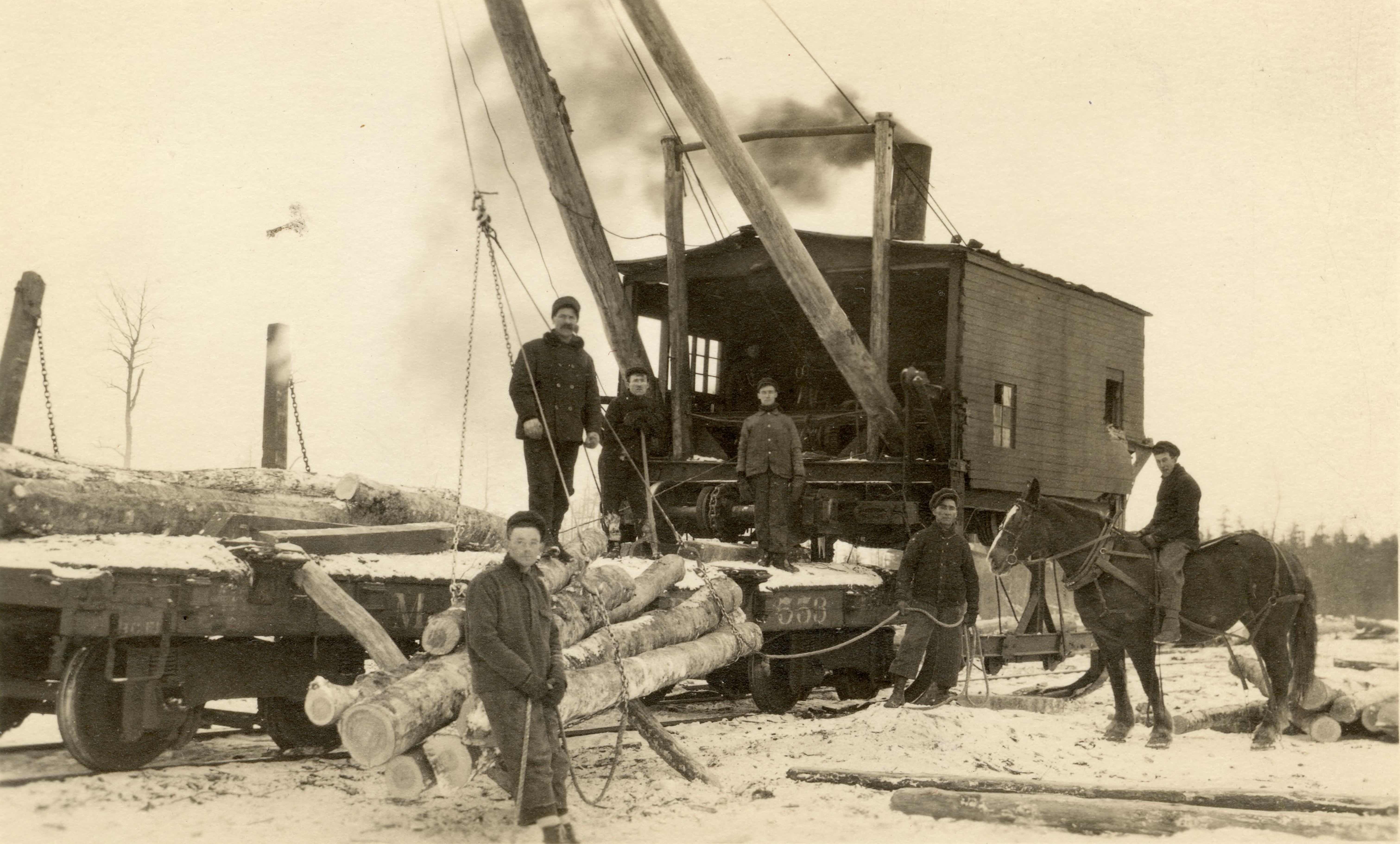 Men stand in front of a lumber train. Another man is on horseback next to them.