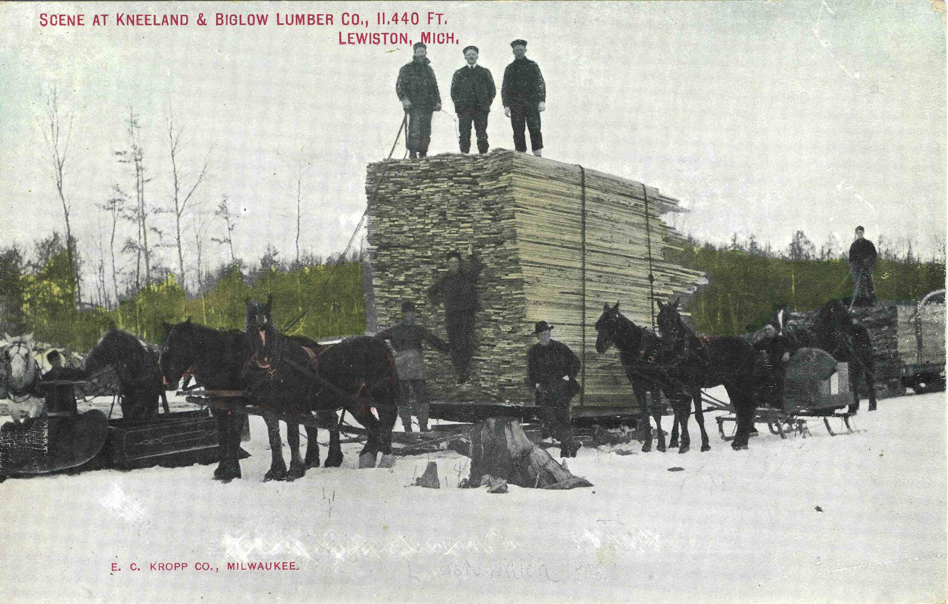 Three men stand on top of a tile pile of lumber being carried by a team of horses during the winter