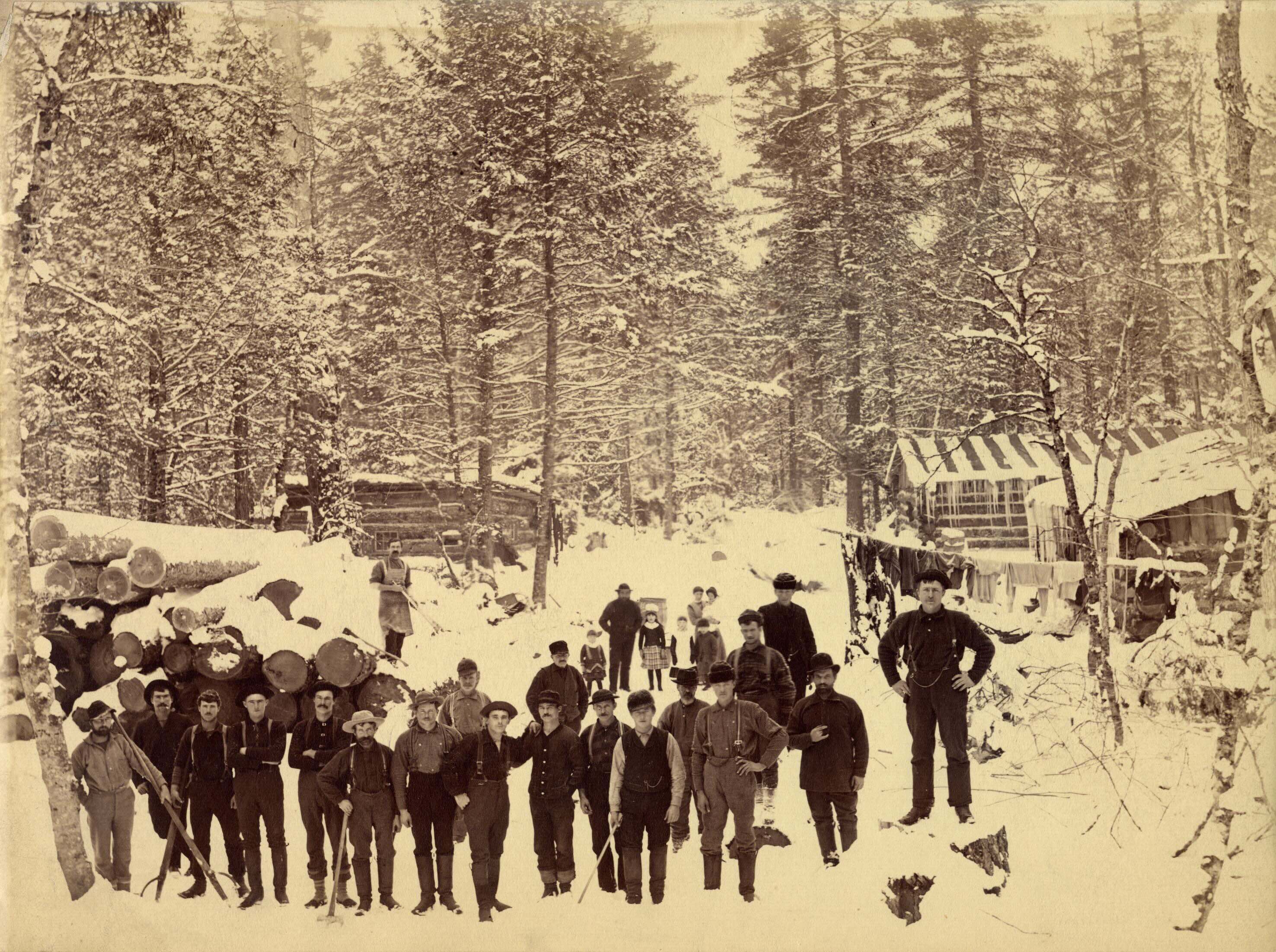 Men and boys stand in front of a lumber camp in the winter