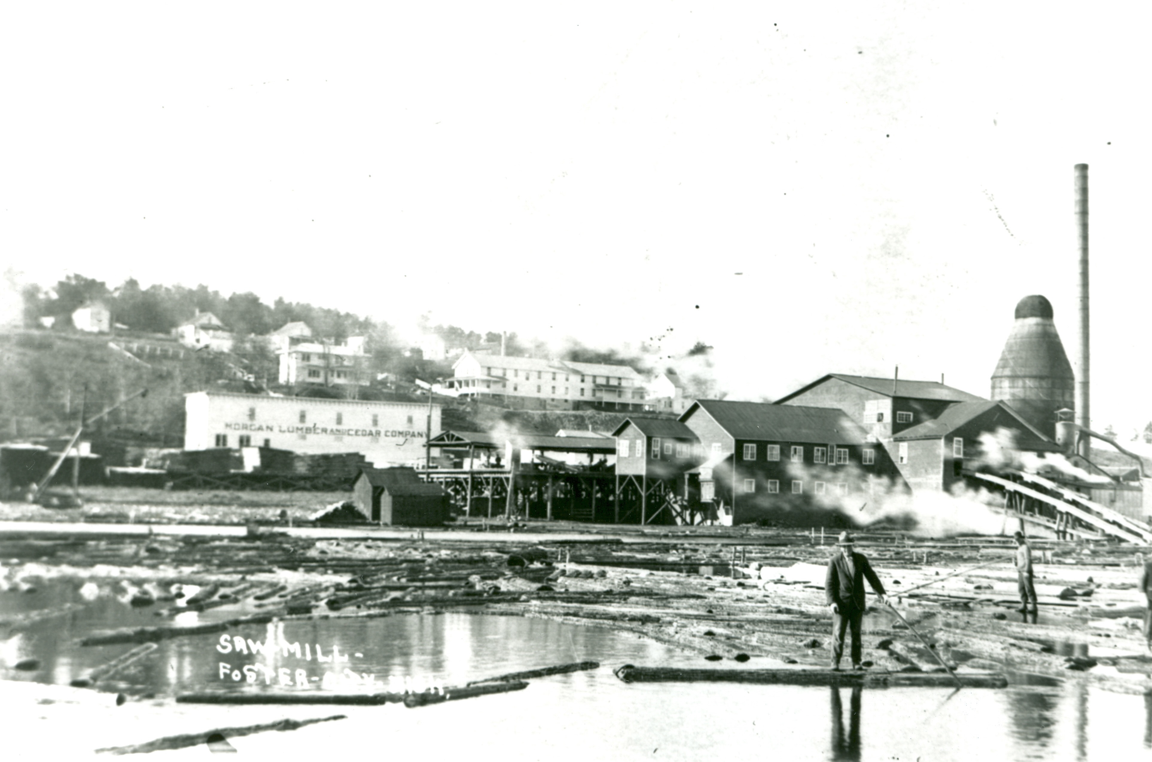 A man in a suit stands on a log in a river with a sawmill in the background