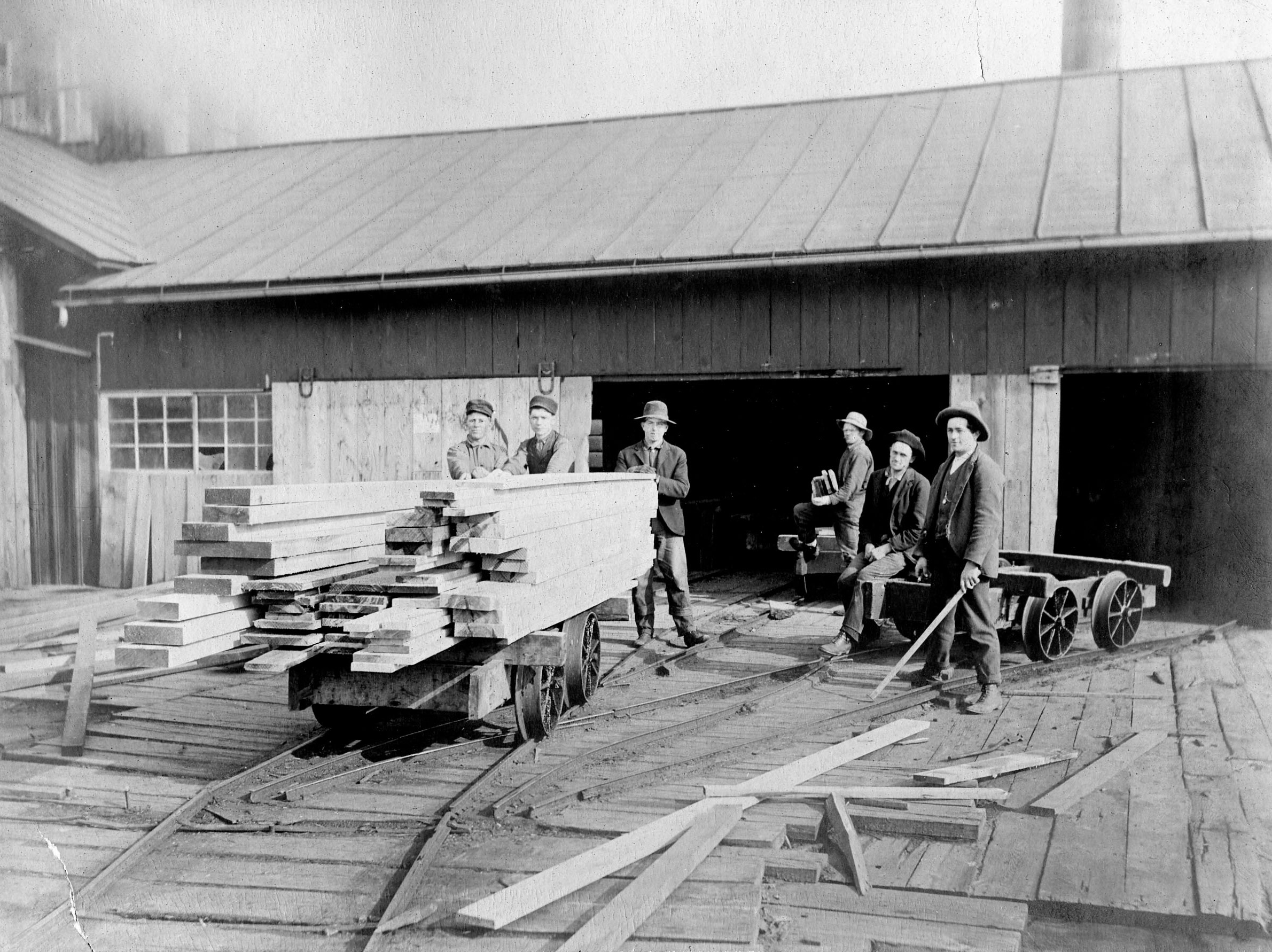 Six men stand next to four wheeled carts stacked with lumber in front of a sawmill