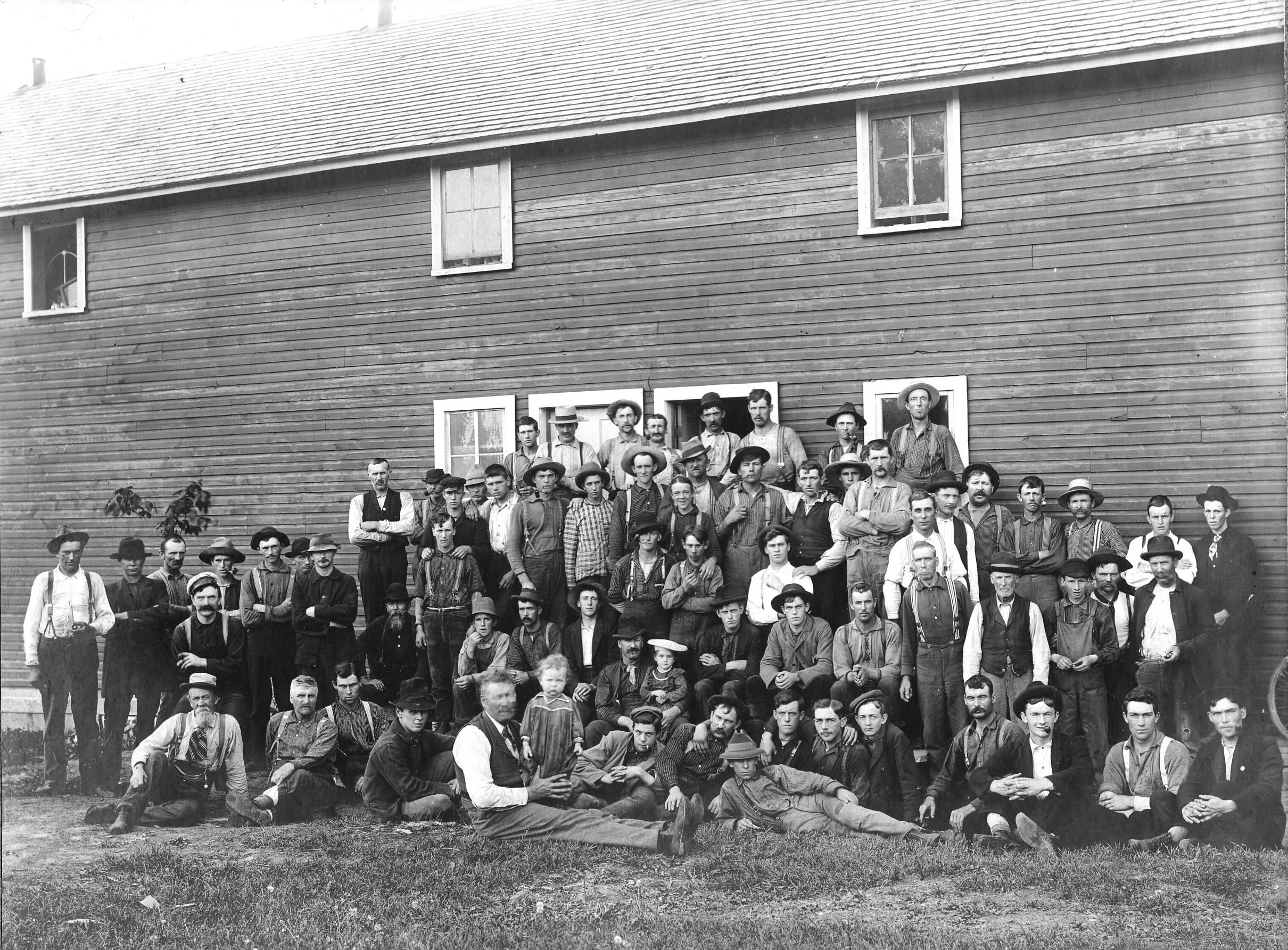 A group of over 50 men stand in front of a wooden building